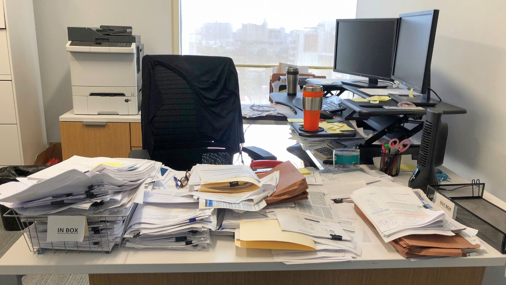 A cluttered office desk with piles of papers and folders covers most of the surface. Dual monitors and office supplies suggest a busy, chaotic work environment.